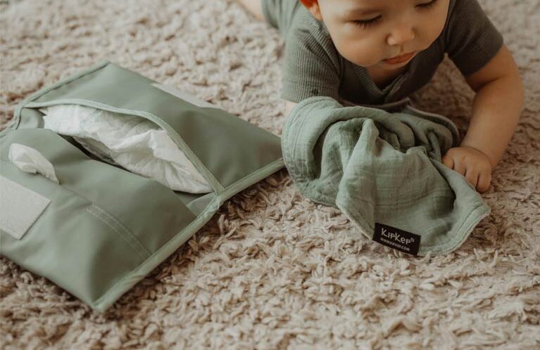 Baby with green muslin cloth and diaper pouch on carpet